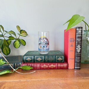 May include: A vintage clear glass featuring Mickey Mouse with the text "Mickey" and "1955". The glass is set on a stack of antique books with green, red, and orange covers. A leafy plant is on the left.