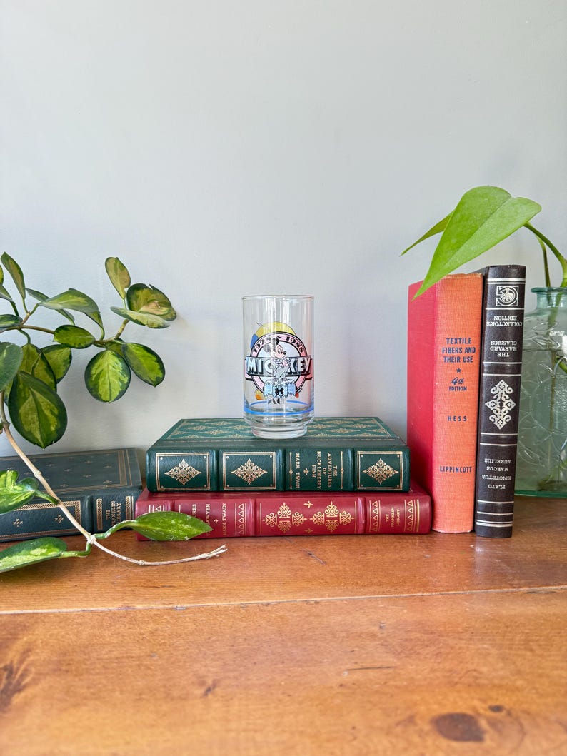 May include: A vintage clear glass featuring Mickey Mouse with the text "Mickey" and "1955". The glass is set on a stack of antique books with green, red, and orange covers. A leafy plant is on the left.