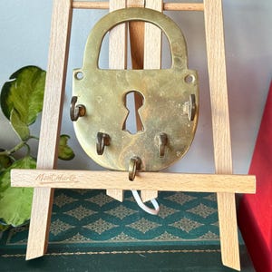 Puede incluir: Un llavero de latón antiguo con forma de candado grande, con cuatro ganchos para llaves. El llavero está montado en un pequeño caballete de madera. El caballete está colocado frente a un libro verde con letras doradas y un libro rojo.