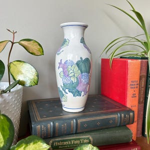 May include: A white ceramic vase with a blue and green floral design. The vase is decorated with a variety of flowers and leaves, including pumpkins, gourds, and other foliage. The vase is sitting on a stack of books.