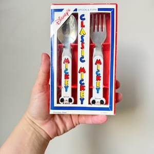 Może przedstawiać: Zestaw łyżki i widelca ze stali nierdzewnej Disney Classic Mickey w prostokątnym pudełku. Białe uchwyty mają napis Mickey czerwonymi, żółtymi i niebieskimi literami z ikoną głowy Myszki Miki u podstawy. Pudełko zawiera napis Disney.