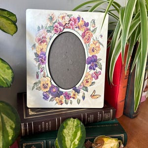 Puede incluir: Un marco de fotos blanco con un dise&ntilde;o floral con pensamientos morados, amarillos y rosas. El marco tiene una abertura ovalada para una foto.