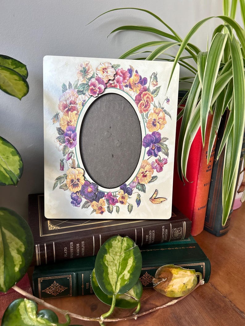 Puede incluir: Un marco de fotos blanco con un dise&ntilde;o floral con pensamientos morados, amarillos y rosas. El marco tiene una abertura ovalada para una foto.
