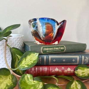 May include: A red, blue, and orange glass bowl with a wavy design. The bowl is sitting on a stack of books with titles like "Andersen's Fairy Tales" and "Little Women".