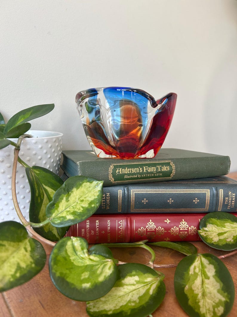 May include: A red, blue, and orange glass bowl with a wavy design. The bowl is sitting on a stack of books with titles like "Andersen's Fairy Tales" and "Little Women".