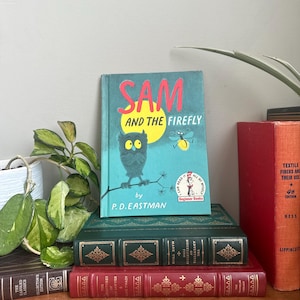 Può includere: Un libro per bambini con copertina rigida intitolato Sam and the Firefly di P.D. Eastman con una copertina color ottanio che raffigura un gufo e una lucciola. Il libro poggia su due libri rilegati in pelle, uno verde scuro e uno rosso.