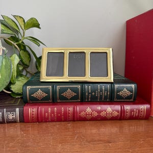 May include: A gold-toned triple photo frame with a dark interior, placed on a stack of vintage books. The books are in shades of green and red, with gold lettering and decorative patterns. The frame has the text "1918" on it.