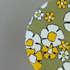 May include: A round, vintage metal tray with a floral pattern. The tray features large white and yellow flowers with black outlines on a green background. The tray is held by a hand.