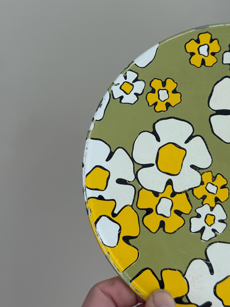 May include: A round, vintage metal tray with a floral pattern. The tray features large white and yellow flowers with black outlines on a green background. The tray is held by a hand.