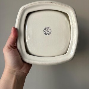 Op de afbeelding: Witte vierkante keramische schaal met een verhoogde rand en een kleine, ronde, zwart-witte stempel op de bodem met de tekst "Made in Japan".