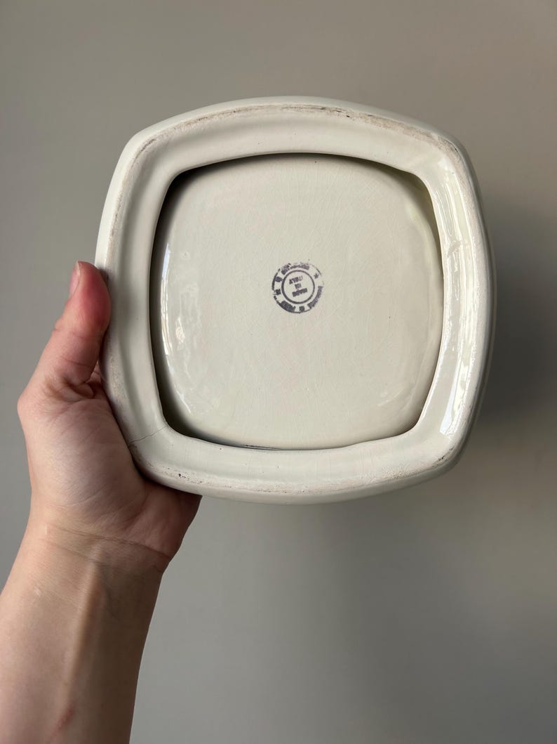 Op de afbeelding: Witte vierkante keramische schaal met een verhoogde rand en een kleine, ronde, zwart-witte stempel op de bodem met de tekst "Made in Japan".