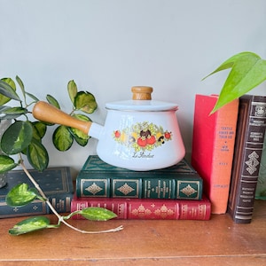 Peut inclure: Pot à fondue en émail blanc avec une poignée et un couvercle en bois, décoré d'un motif de fruits et de légumes. Les mots "La Fondue" sont imprimés sur le pot. Le pot est posé sur une pile de livres anciens.