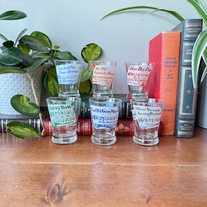 May include: A collection of six vintage shot glasses, each printed with song lyrics and musical notation. The glasses display songs such as "You Tell Me Your Dream" and "The Old Gray Mare." They are arranged on a wooden surface, with books and plants visible in the background.