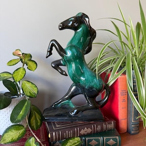 Op de afbeelding: Een groen en zwart keramiek paardenbeeldje met een geglazuurde afwerking. Het paard staat op zijn achterpoten met zijn voorpoten omhoog. Het beeldje staat op een stapel boeken.