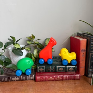 Può includere: Tre veicoli giocattolo colorati: una tartaruga verde, una giraffa rossa e un'anatra gialla. Ogni giocattolo ha ruote blu. I giocattoli sono disposti su una pila di libri d'epoca, con vegetazione sullo sfondo.