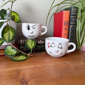 May include: Two white ceramic mugs with handles and 3D noses. One mug features a black winking face with a smirk. The other mug displays a smiling face with closed eyes and small red heart accents near one eye. Both mugs have a smooth finish.