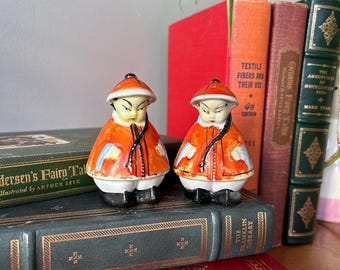 May include: Two small ceramic figurines of people wearing traditional Chinese clothing. The figurines are painted in orange, white, and black. They are sitting on a stack of books.