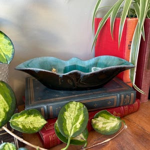 以下が含まれることがあります： 波打つ縁のある、緑がかった青と黒のセラミックボウル。ボウルは、緑の植物が前景にある本の積み重ねの上に置かれています。