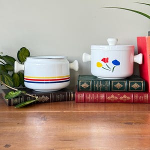 May include: Two white enamelware pots with handles and lids. One pot has a design of red, yellow, and blue stripes. The other pot has a floral design of red, yellow, and blue flowers. Both pots are sitting on a stack of books.