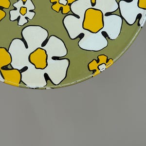 May include: Close-up of a round, vintage metal tray with a floral pattern. The flowers are white with yellow centers and black outlines, set against a muted green background. The tray's edge is yellow.
