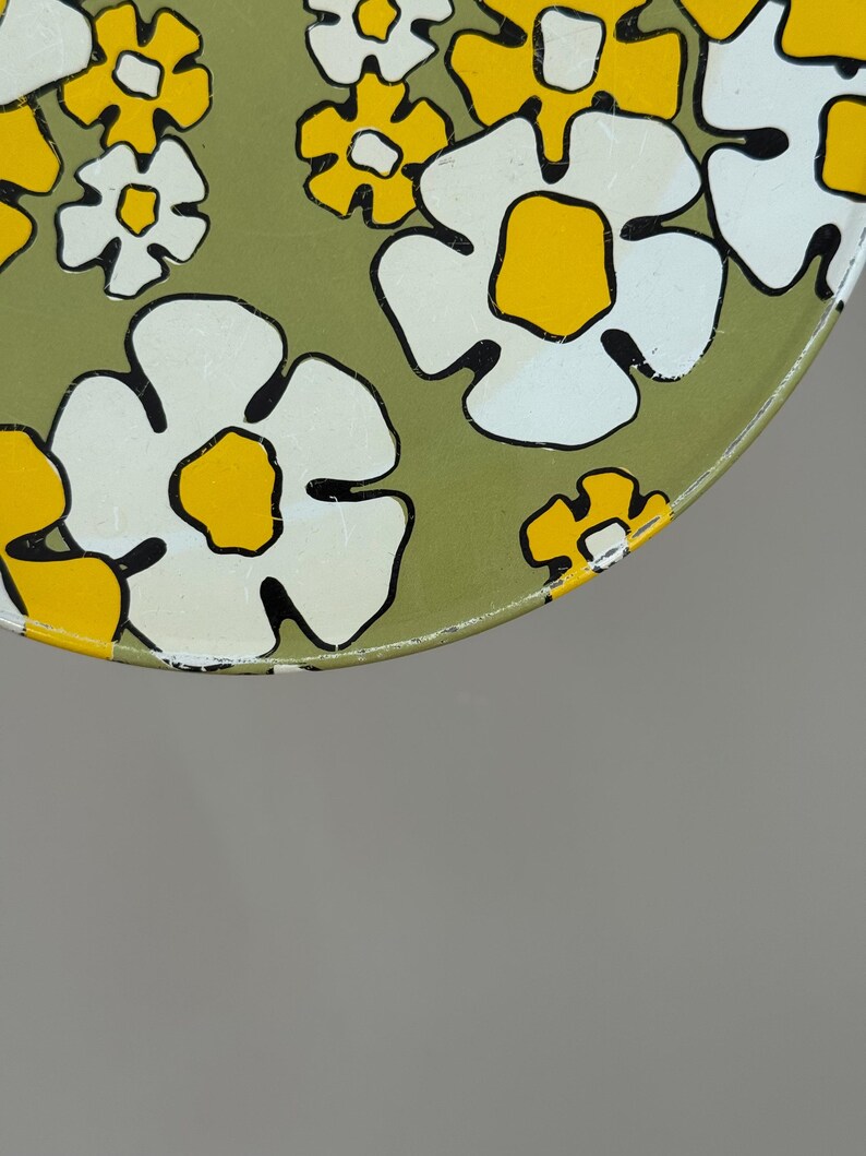 May include: Close-up of a round, vintage metal tray with a floral pattern. The flowers are white with yellow centers and black outlines, set against a muted green background. The tray's edge is yellow.