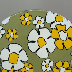 May include: Close-up of a section of a round, olive green surface with a repeating floral pattern. The flowers are white and yellow with black outlines. The design is reminiscent of a retro style.