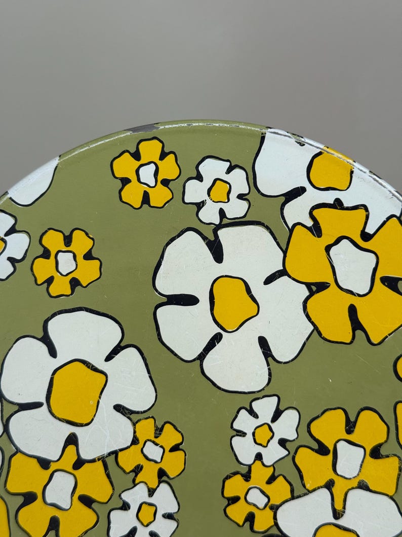 May include: Close-up of a section of a round, olive green surface with a repeating floral pattern. The flowers are white and yellow with black outlines. The design is reminiscent of a retro style.