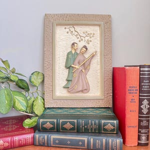 May include: A framed relief sculpture depicting two figures in traditional Asian attire, one holding a staff. The artwork is set against a textured background and framed in a light brown color. The sculpture rests on a stack of vintage books with various colors and titles.