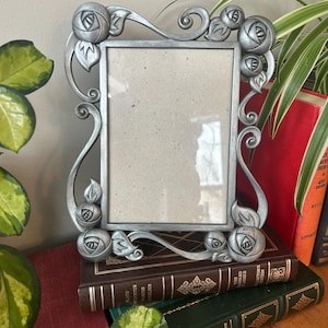 Könnte beinhalten: Ein silberfarbener Bilderrahmen mit einem dekorativen Design aus Rosen und Schnörkeln. Der Rahmen steht auf einem Stapel Bücher.