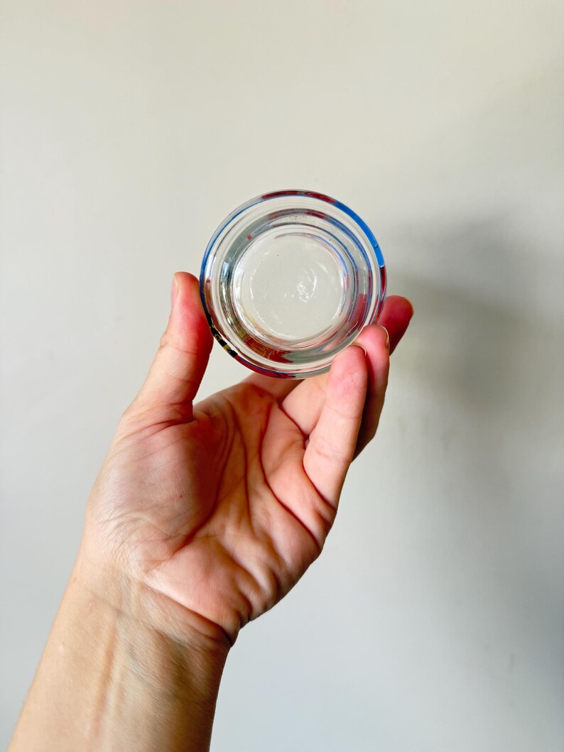 May include: A clear, round glass object with a colorful rim of blue, red, and green. The object is held in a hand against a plain background. The glass appears to be a small container or lid.