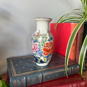 May include: A small white ceramic vase with a floral design. The vase features a delicate, hand-painted design with pink, red, blue, and green flowers. The vase is sitting on top of a stack of books.