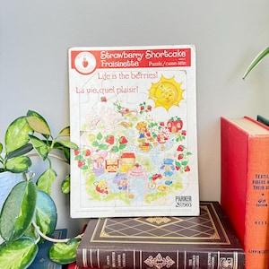 May include: A vintage Strawberry Shortcake puzzle with the text "Life is the berries!" and "La vie, quel plaisir!". The puzzle features colourful illustrations of Strawberry Shortcake characters and strawberries. The puzzle is on a stack of books.