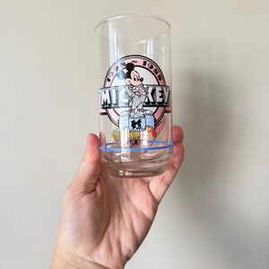 May include: Clear glass tumbler featuring a Mickey Mouse graphic. The design includes the text "MICKEY" in a banner, with the year "1988" in a circle. A blue stripe encircles the base of the glass.