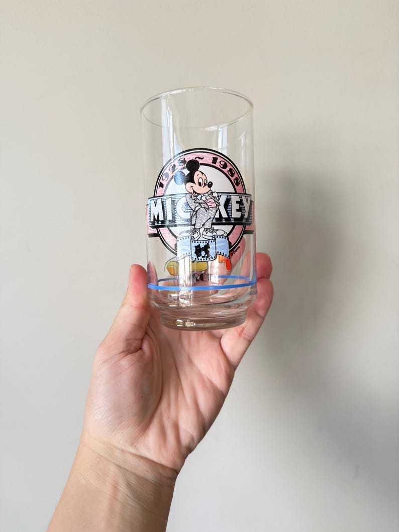 May include: Clear glass tumbler featuring a Mickey Mouse graphic. The design includes the text "MICKEY" in a banner, with the year "1988" in a circle. A blue stripe encircles the base of the glass.