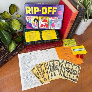 Könnte beinhalten: Ein Vintage-Brettspiel mit dem Titel "RIP-OFF" mit farbenfrohen Illustrationen und Text. Das Spiel beinhaltet Spielgeld, Anleitungsblätter und Spielkarten. Die Box ist lila und zeigt Cartoon-Bilder. Weitere Artikel sind Bücher und Pflanzen.