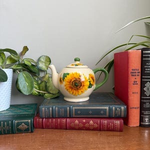 May include: A cream-colored ceramic teapot with a sunflower design, green handle and lid. The teapot sits atop a stack of vintage books with red, green, and blue covers. A potted plant is visible on the left.