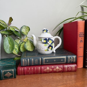 以下が含まれることがあります： 青と黄色の浮き彫りの花柄が施された白い陶器のティーポットが、古書の上に置かれています。ティーポットは、曲がったハンドルと注ぎ口が付いています。本は、緑と赤など、さまざまな色で製本されています。
