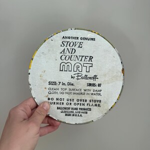 May include: A round stove and counter mat with the text "STOVE AND COUNTER MAT" in black block letters. The mat is 7 inches in diameter and has a yellow and white floral border. The text "ANOTHER GENUINE" is at the top.