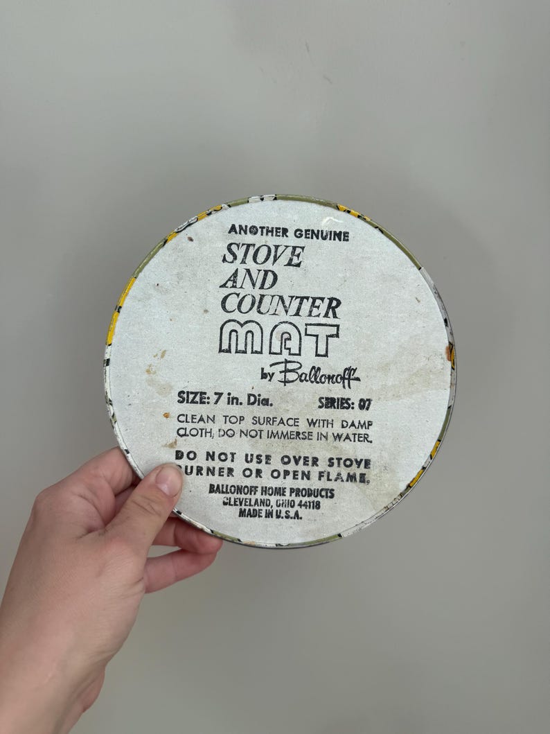 May include: A round stove and counter mat with the text "STOVE AND COUNTER MAT" in black block letters. The mat is 7 inches in diameter and has a yellow and white floral border. The text "ANOTHER GENUINE" is at the top.