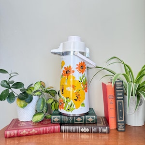 May include: A vintage white and floral thermos with a white handle and lid. The thermos features large yellow and orange flowers with green leaves. The thermos is sitting on a stack of books.
