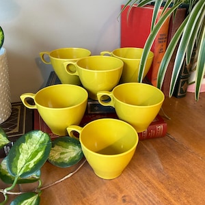 May include: Six vibrant yellow teacups with handles are displayed on a wooden surface. The cups have a classic design and are made of a solid, opaque material. The background includes books and greenery.