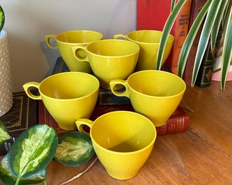 May include: Six bright yellow teacups with handles are arranged on a wooden surface. The cups have a classic design and are made of a solid, opaque material. The background includes books and greenery.