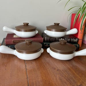 May include: Four white ceramic ramekins with brown lids and handles. The ramekins are arranged on a wooden surface with books stacked behind them.