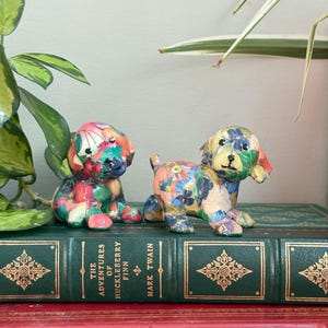May include: Two small dog figurines decorated with a colourful floral decoupage pattern. One dog is in a sitting position, while the other is standing. Both figures feature multi-coloured petals and leaves in shades of pink, blue, green, and yellow.