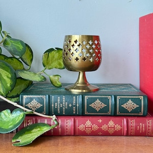May include: A vintage brass candle holder with a pierced design, sitting atop two antique books. The books are bound in green and red leather, with gold lettering and decorative patterns. The candle holder has a pedestal base and a cup-shaped top with cross-shaped cutouts.
