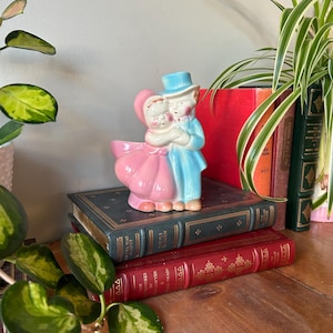 Puede incluir: Un macetero de cerámica vintage con forma de pareja tomándose de la mano. La mujer lleva un vestido rosa y el hombre un traje azul. El macetero está sobre una pila de libros.
