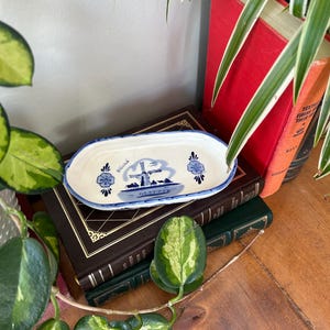 Può includere: Un piatto ovale in ceramica con base bianca e bordo blu. L'interno presenta un'illustrazione dipinta in blu di un mulino a vento, motivi floreali e la parola Holland scritta in corsivo. Il piatto ha una finitura liscia e smaltata.