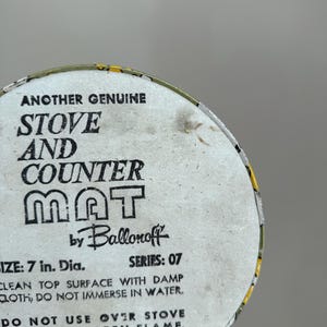 May include: A round stove and counter mat with the text "ANOTHER GENUINE STOVE AND COUNTER MAT" by Ballonoff. The mat is 7 in. in diameter. The text also includes cleaning instructions and a warning about open flames.