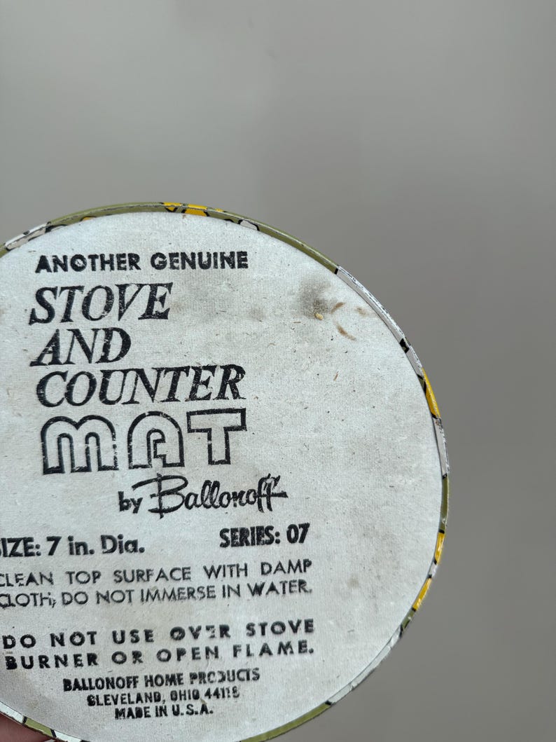 May include: A round stove and counter mat with the text "ANOTHER GENUINE STOVE AND COUNTER MAT" by Ballonoff. The mat is 7 in. in diameter. The text also includes cleaning instructions and a warning about open flames.