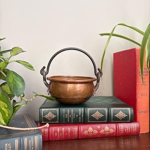 May include: A small, round copper pot with a dark metal swing handle attached to two side loops. The pot has a smooth, metallic finish and sits atop a stack of three hardcover books, including The Adventures of Huckleberry Finn and Tom Sawyer.